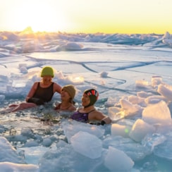 Three people are submerged up to their shoulders in water with broken ice covering the surface.